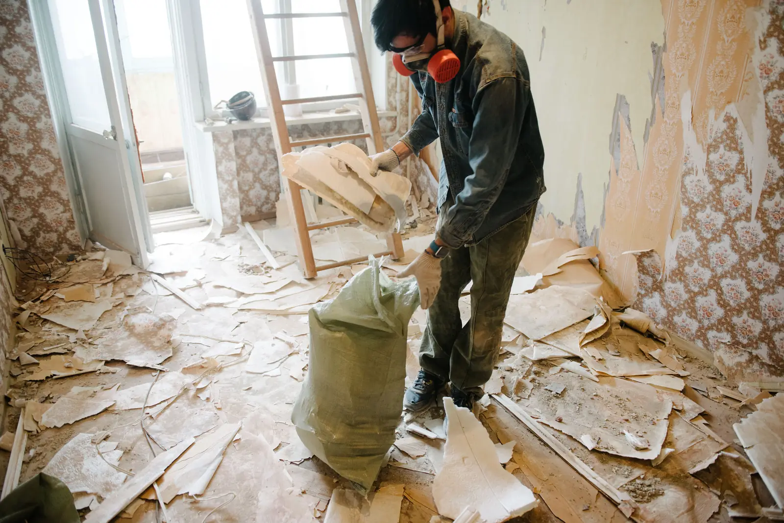 Worker in a respirator bagging torn-out drywall and wallpaper during a demolition cleanup